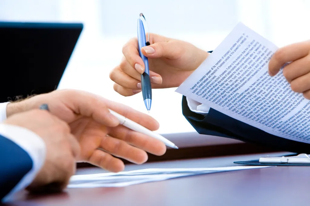 Two people holding pens, pointing to papers on a desk.