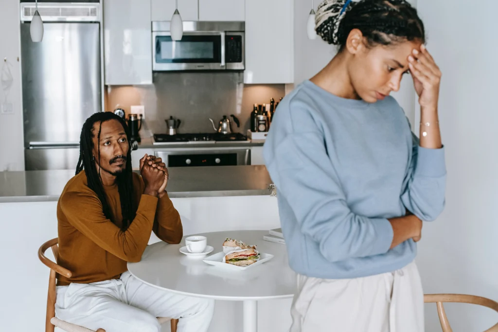 A couple in their kitchen, one person sitting at the table and the other standing holding her head.